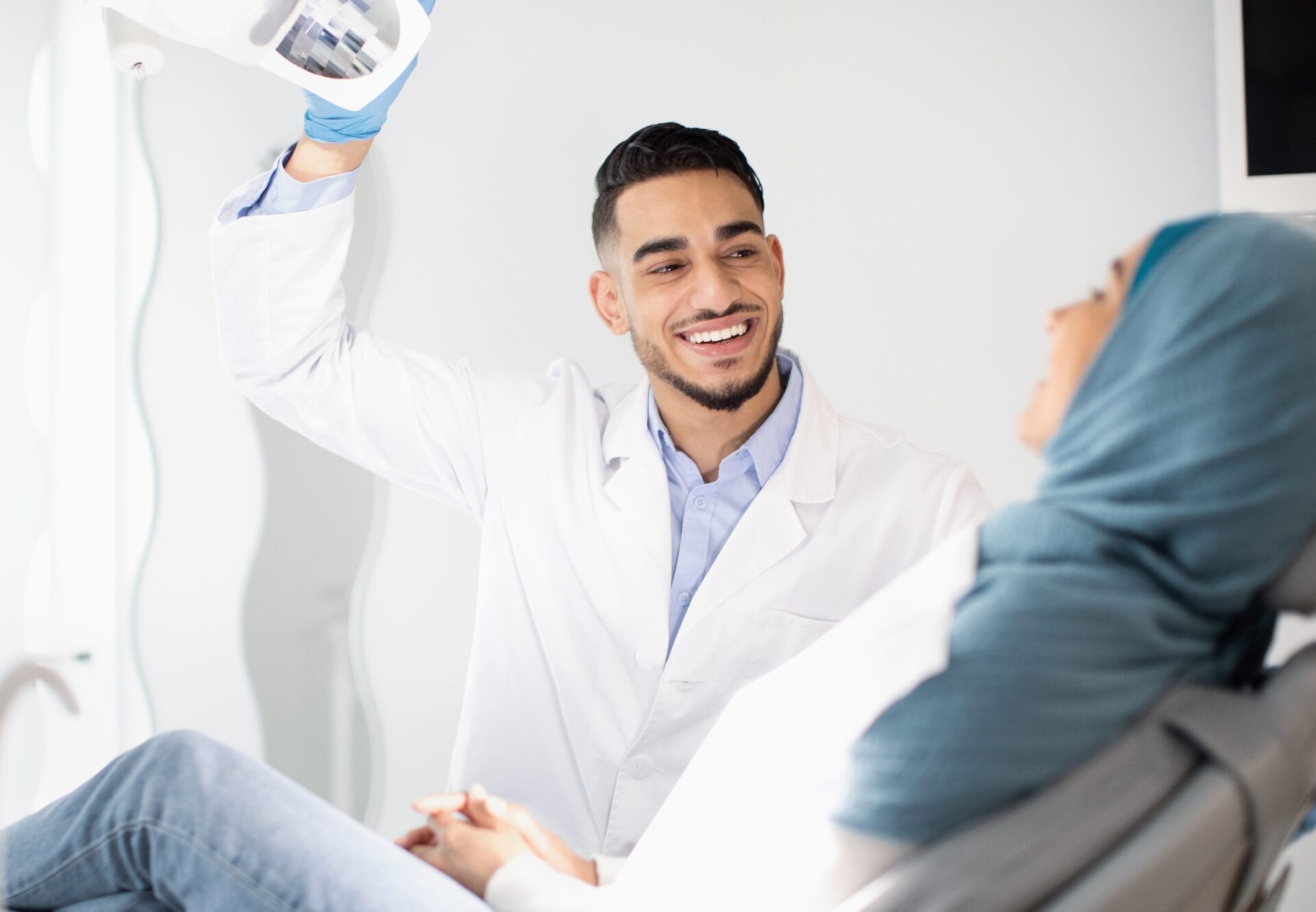 Dental Treatment. Portrait Of Middle Eastern Dentist Doctor Having Check Up With Muslim Female Patient In Modern Clinic, Friendly Arab Stomatologist Man Turning On Lamp And Smiling, Close Up