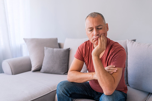 Man sitting on the couch and holding his jaw in pain, needing toothache treatment at Dens Dental Studio in Uptown Chicago, IL.